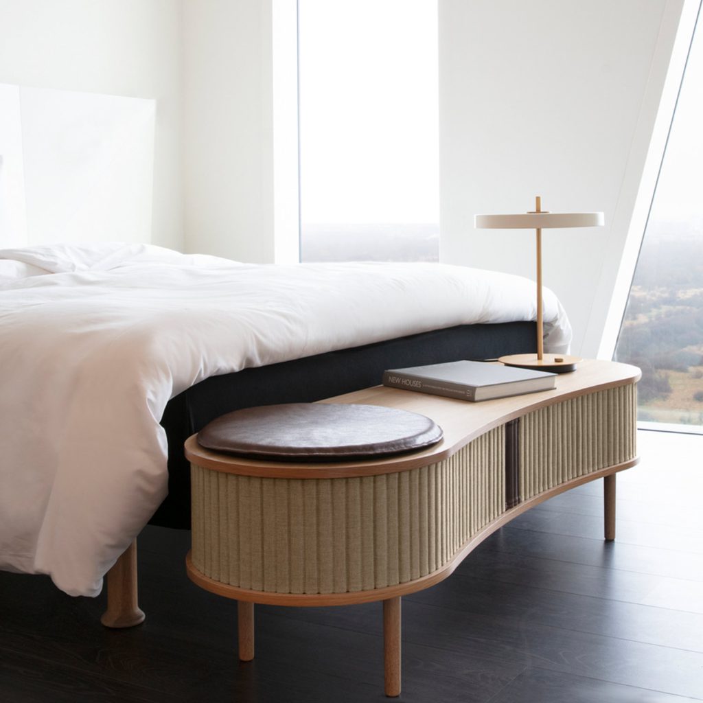 Natürlich wohnen: wirklich coole Ideen mit Holz, Glas & Erdtönen 1 Lowboard aus Holz und Textil in organischer Form mit Leuchte, Buch und rundem Sitzpolster vor einem Doppelbett.