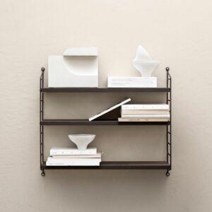 Das dunkle String Furniture Pocket Regal an einer hellen Wand mit Büchern und ausgewählten Objekten.