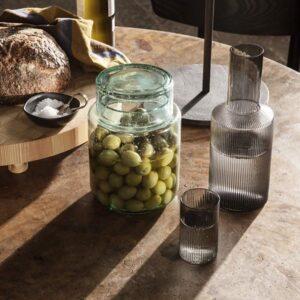 Ferm Living Ripple Glas-Karaffe und ein Ripple Verrines Glas – beide in Grau – auf einem Tisch neben Brot und Oliven.